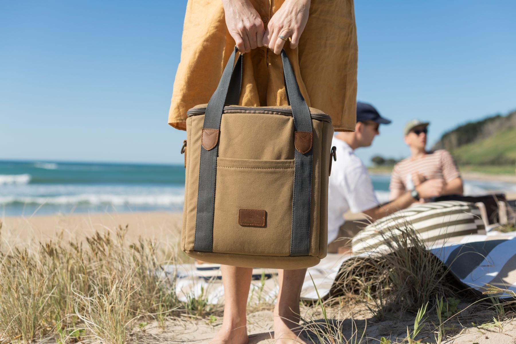 A lady at the beach holding the Slowlife Collection Olive Cooler Bag. This has a front pocket, leather detailing and a long shoulder strap included.