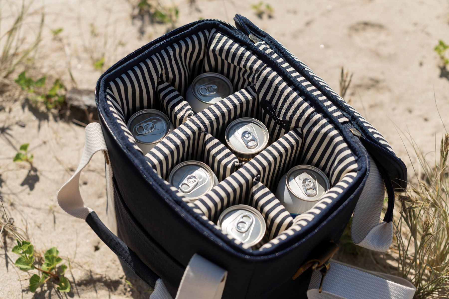 The Slowlife Collection Navy cooler bag on the beach and open. Shows navy and cream stripe interior fill with 12 cans of beer.