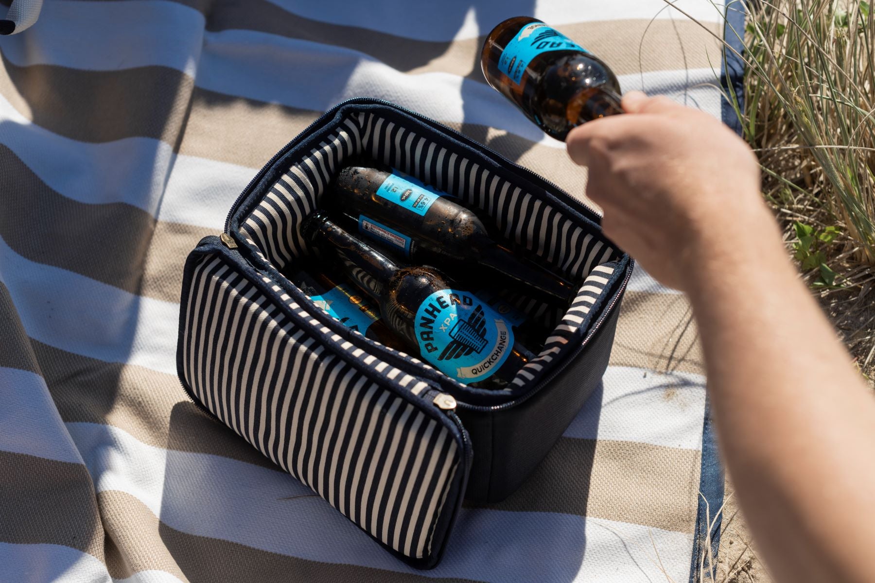 The Navy Slowlife Collection Lunch Bag filled with 6 x panhead bottles of beer. The Lunch Bag is sitting on the Slowlife Collection rug. The Lunch Bag has navy and cream striped interior. There is a hand pulling a beer out of the Lunch Bag.