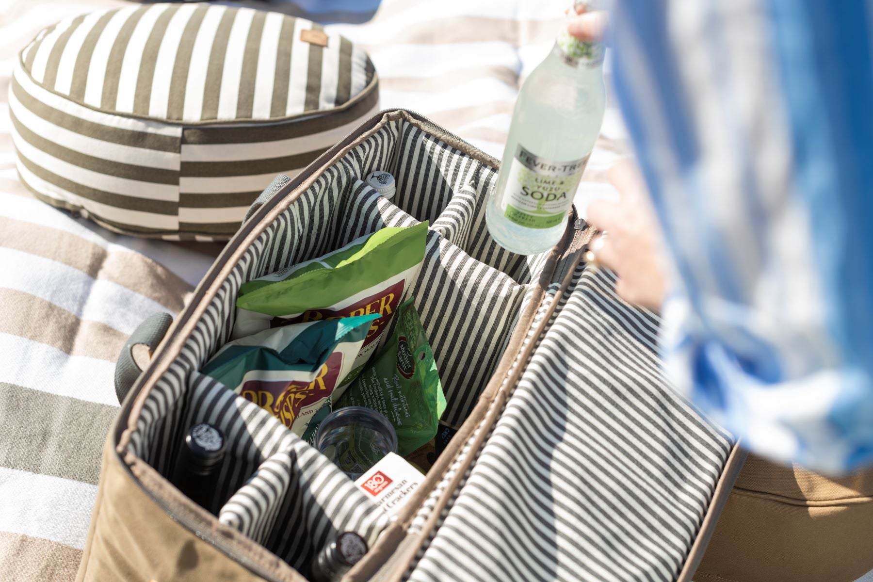 The Olive Slowlife Collection Picnic Bag sitting on the Slowlife Collection Picnic Rug. The bag is open showing the Olive and cream lining. The Picnic bag is full with chips, drinks and snacks. The Slowlife Collection Olive strip cushion is sitting in the background.