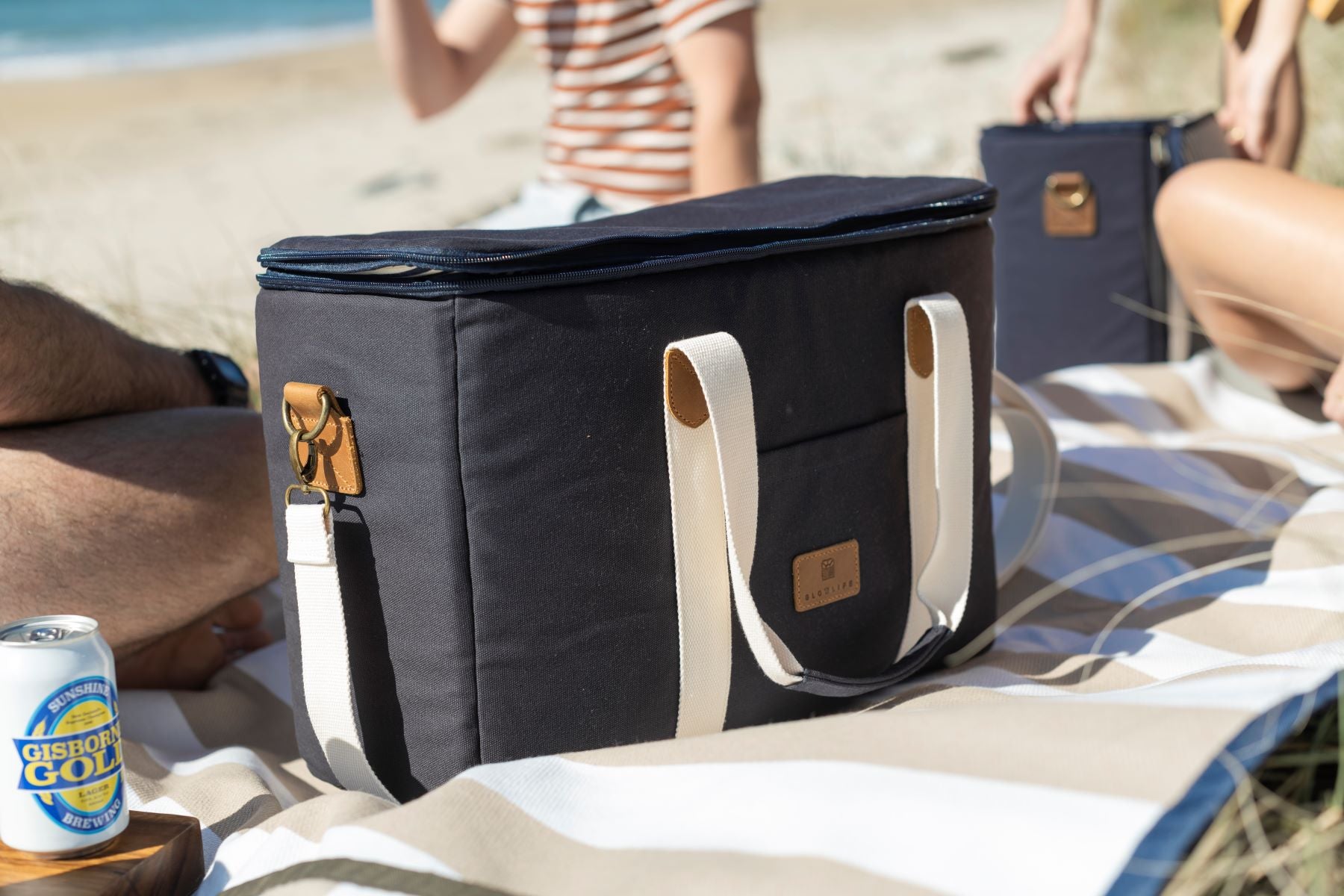 The Navy Slowlife Collection Picnic Bag sitting on the Slowlife Collection rug. Its at the beach and there are people in the background relaxing.