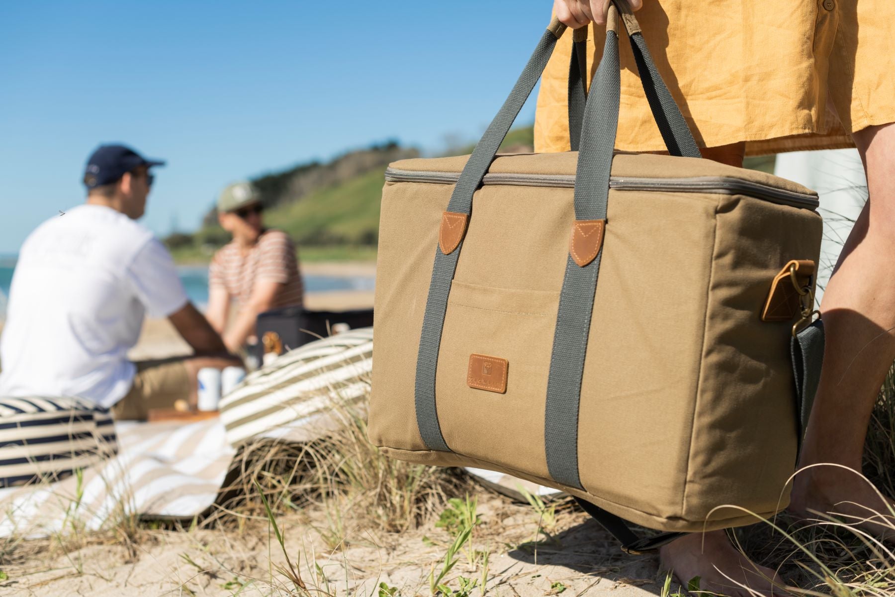 A lady holding the Olive Slowlife Collection Picnic Bag at the beach with people having a picnic in the background.