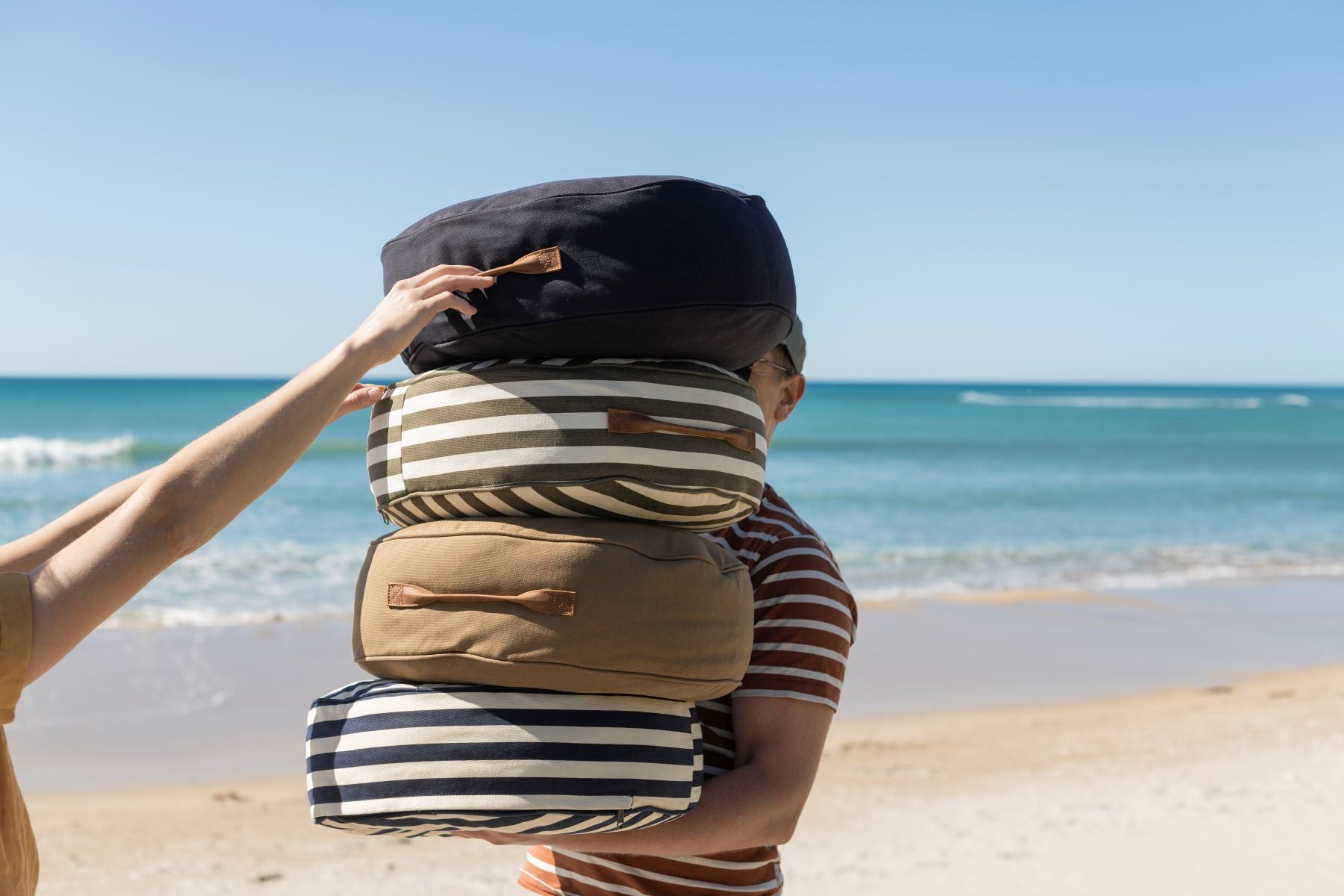 A man holding a stack of Slowlife Collection cushions at the beach (navy, olive striped, olive and navy striped.  You can see a ladies hand grabbing the top cushion from the pile.
