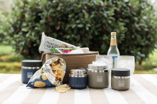 The whole Slowlife Collection range, featuring Navy and Olive in both 20oz and 8oz. Sitting on the Slowlife Collection picnic rug with the Slowlife Collection Olive Lunch Bag with a couple of drinks and a bag of chips.