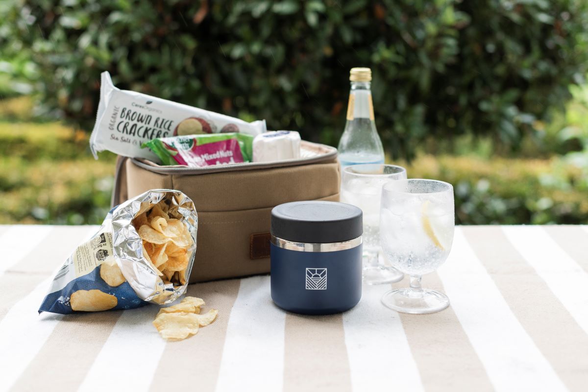 The Navy Slowlife Collection 20oz Flask on the Slowlife Collection Picnic Rug with the Olive Slowlife Collection Lunch Bag and a couple of drinks and a bag of chips. The Navy Flask has the black lid on. 