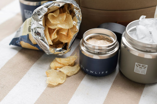 The Navy 8oz Slowlife Collection Flask filled with dip and Olive 20oz Slowlife Collection Flask filled with ice sitting on the Slowlife Collection Picnic Rug beside a bag of open chips. 