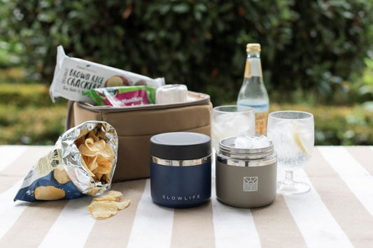 The Navy & Olive Slowlife Collection 20oz Flask on the Slowlife Collection picnic rug with the Olive Slowlife Collection Lunch Bag and a couple of drinks and a bag of chips. The Navy Flask has the black lid on and the Olive Flask is full of ice.