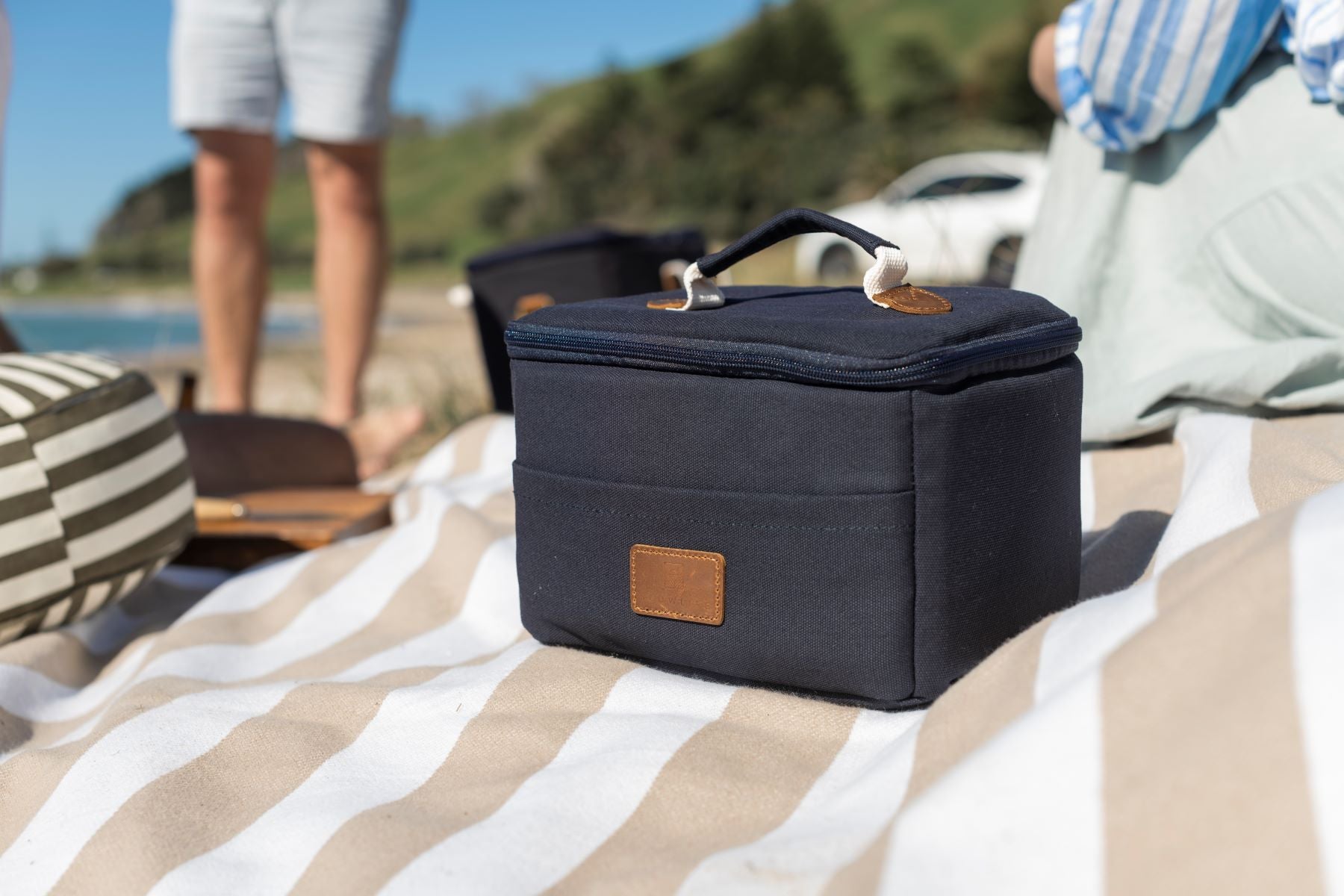 The Navy Slowlife Collection Lunch Bag sitting on the Slowlife Collection Picnic Rug at the beach.