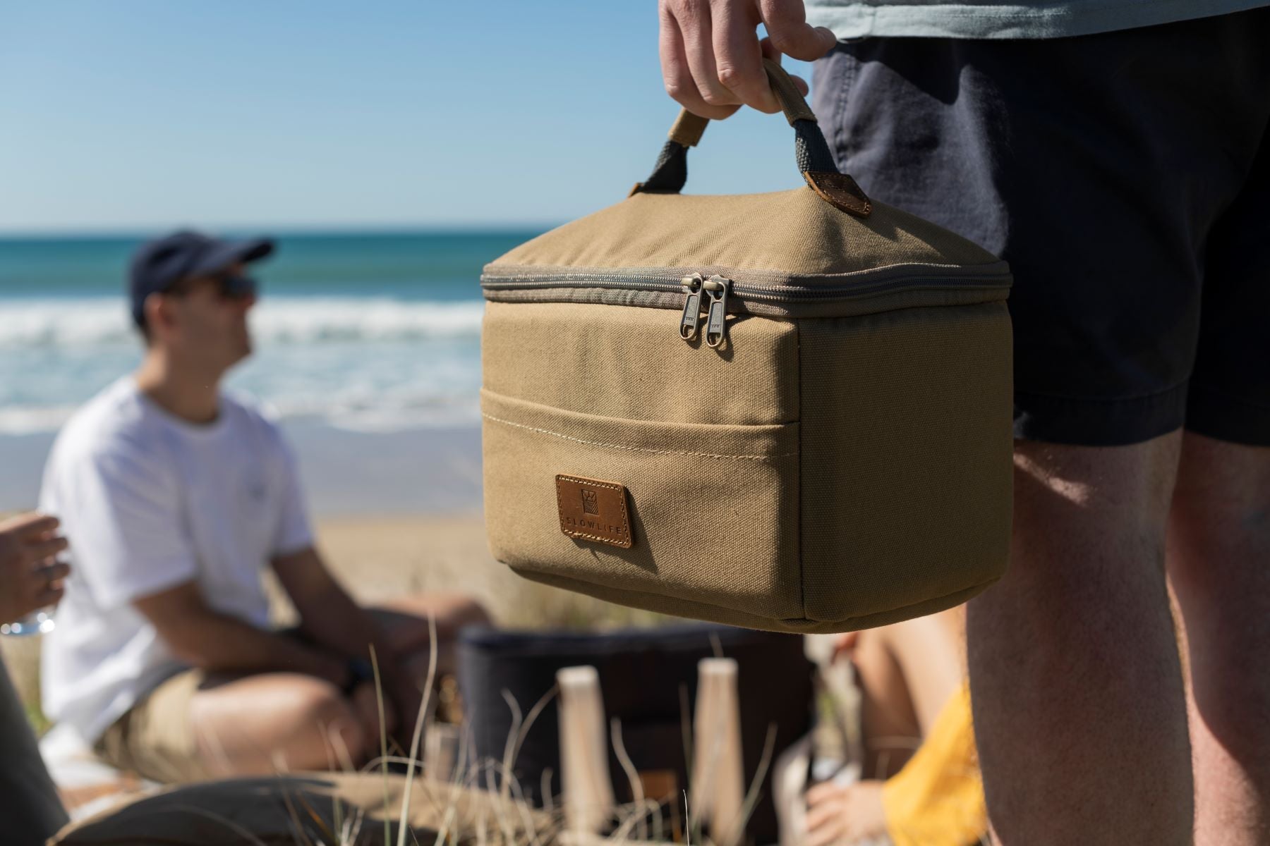 The Olive Slowlife Collection Lunch bag. Being held by the handle and taken at the beach. 