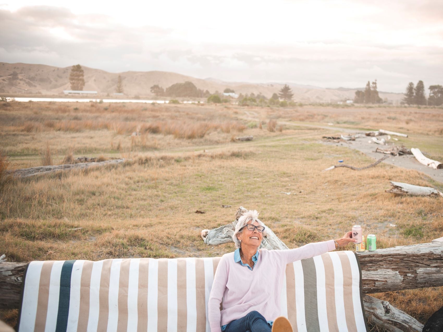 A lady having a drink and smiling, sitting on the Slowlife Collection Picnic Rug which is spread out over a log in the grass. 