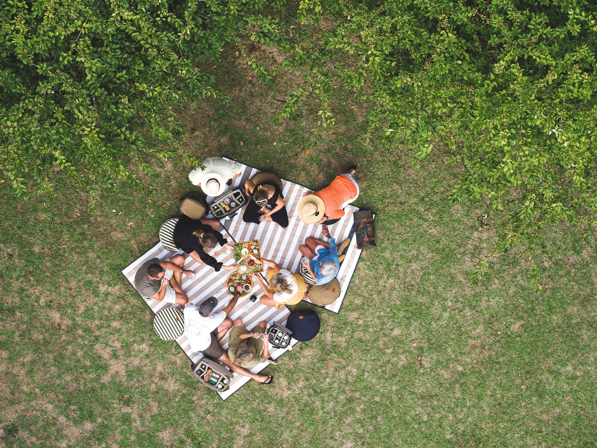 A drone photo looking down on a group of people sitting on the Slowlife Collection Picnic Rugs having a picnic. There are other Slowlife Collection items from the range on the rug too. 