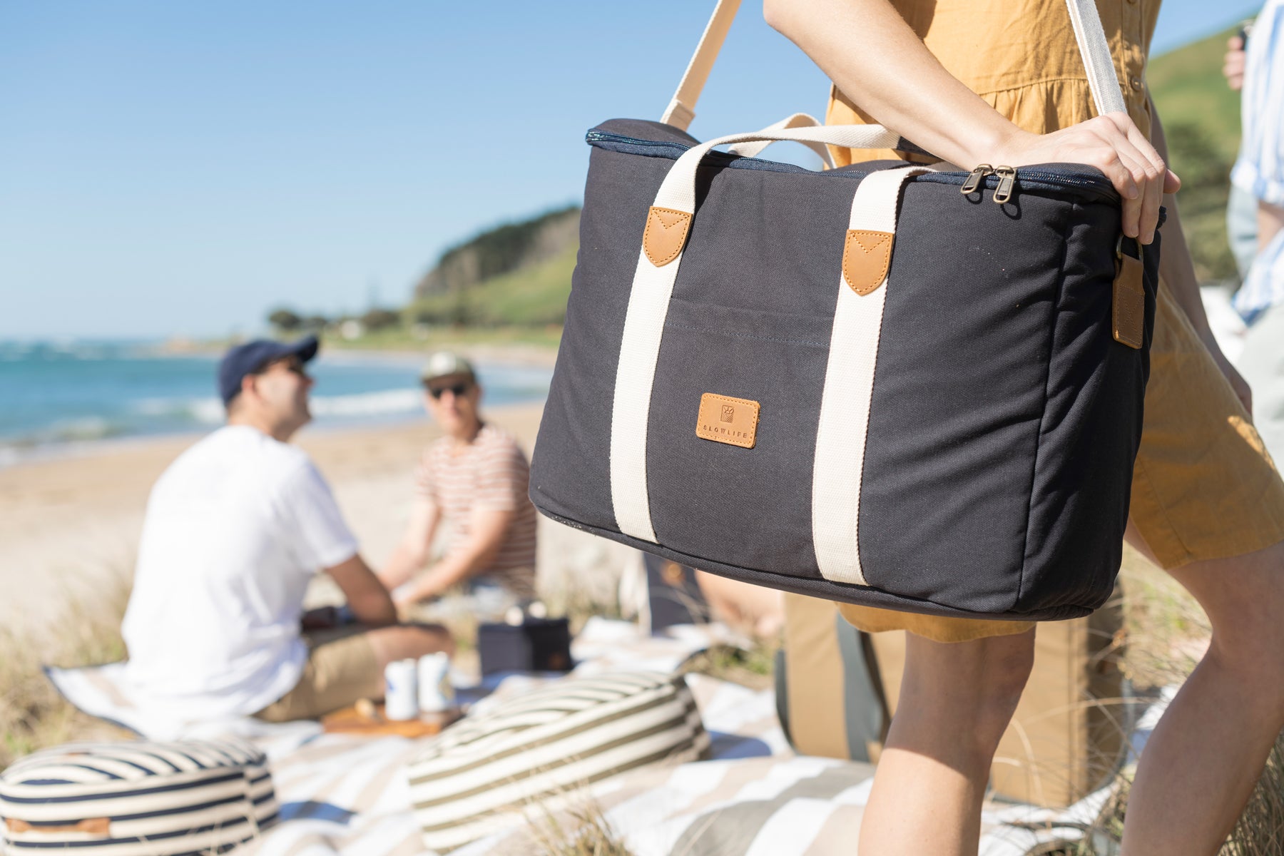A lady standing holding the Navy Slowlife Collection Picnic Bag. She is at the beach at the picnic and has the bag slung over her shoulder. 