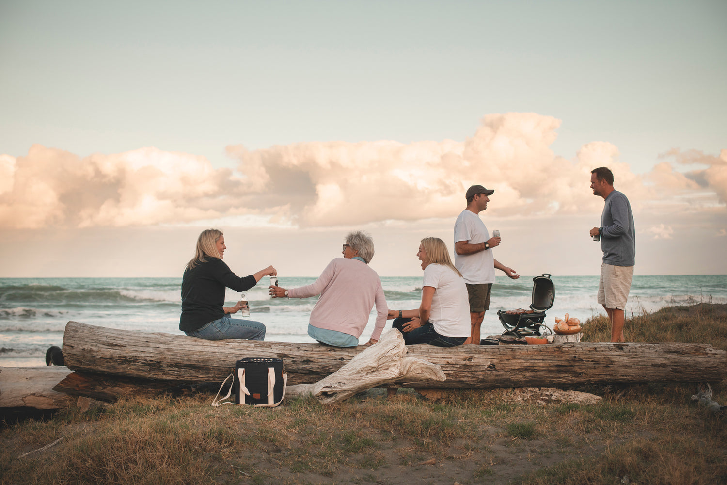 A group of people at the beach at dusk having a BBQ. Three women are sitting talking on a log and there is a Navy Slowlife Collection Cooler Bag in front of them. 