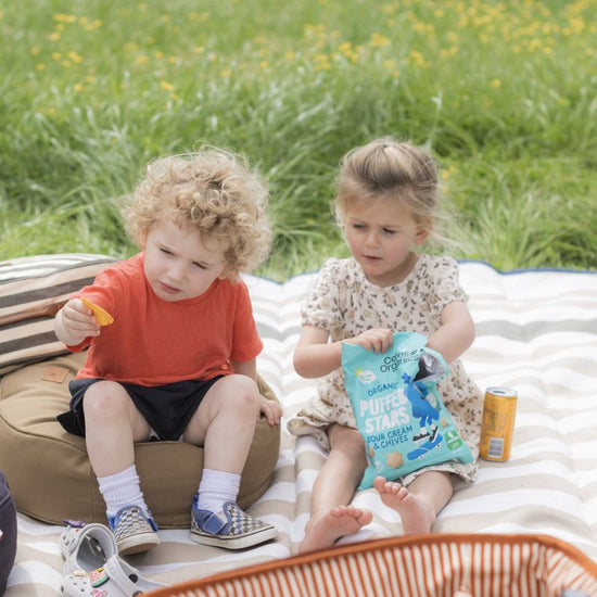 Two children on a striped picnic blanket, snacking outdoors.