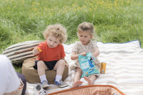 Two children on a striped picnic blanket, snacking outdoors.