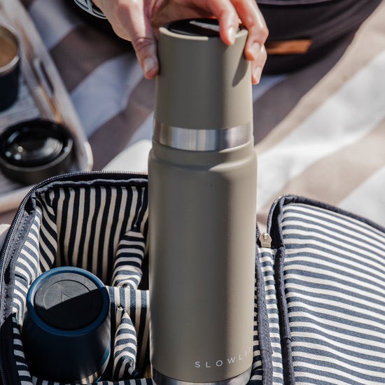 Person holding a olive thermos with Slowlife branding, surrounded by coffee-making equipment on a striped fabric background.