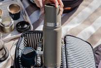 Person holding a olive thermos with Slowlife branding, surrounded by coffee-making equipment on a striped fabric background.