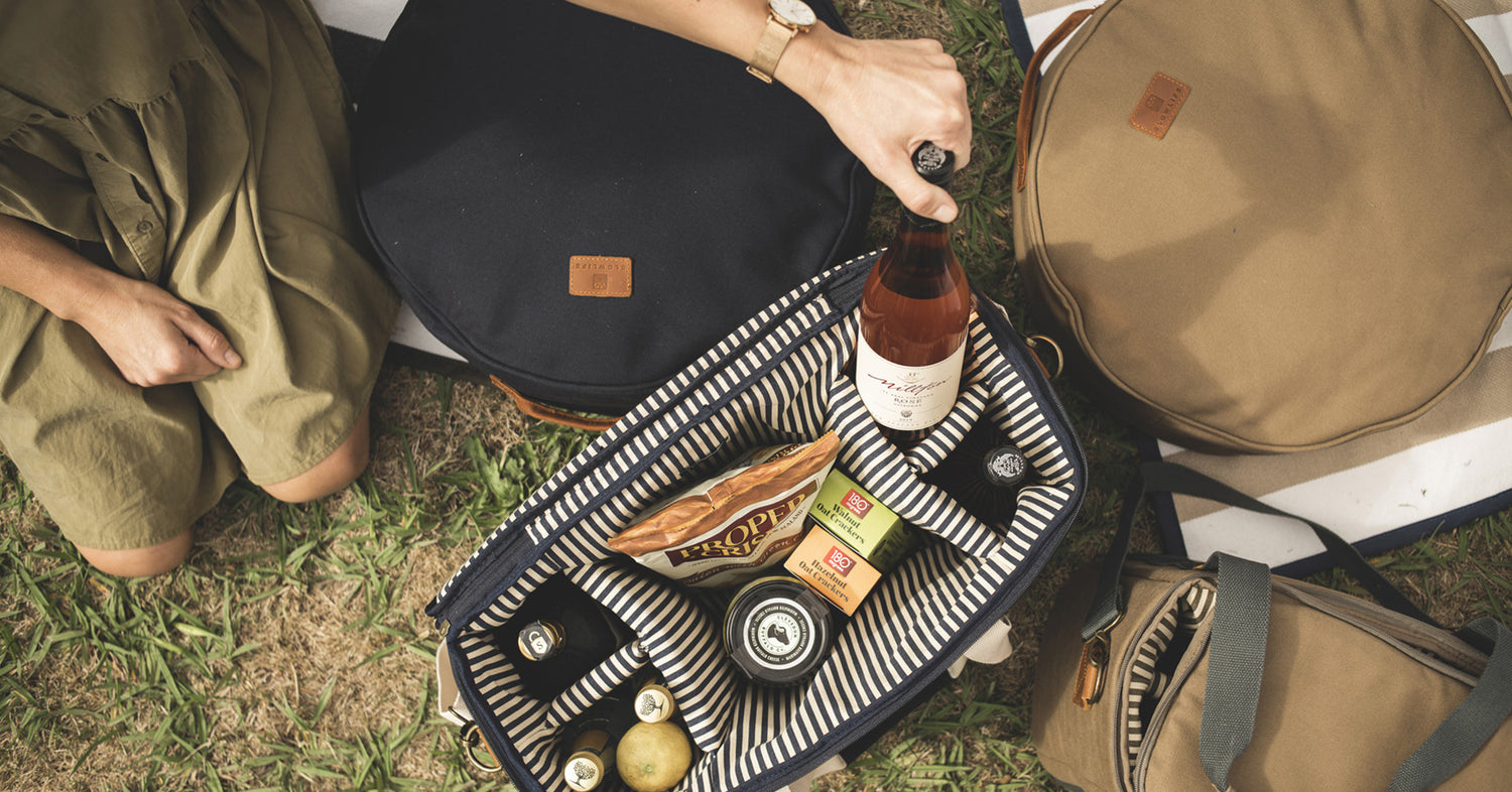 A birds eye view of the lady pulling a bottle of wine out of the Navy Slowlife Collection Picnic Bag. There are also Slowlife Collection Picnic Cushions in the photo as well as a Cooler Bag. The Picnic Bag is packed with wine, chips, crackers and other snacks and beverages. 